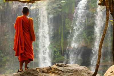 Rear view of buddhist monk standing in front of waterfall