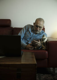 Portrait of adult man lying down on sofa at home and using cellphone with laptop on table