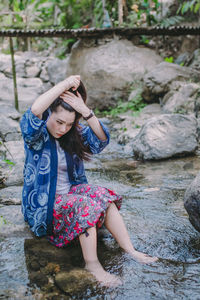 Woman tying hair while sitting on rock amidst lake