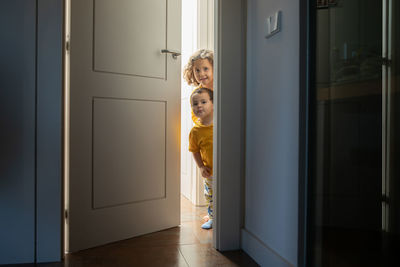 Charming brother and sister peeping out from door in modern apartment while having fun and looking away