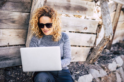 Woman using laptop while sitting against wood