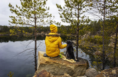 Young woman in yefrom back with fluffy dog sitting on rocky lake near pine trees. travel with pets