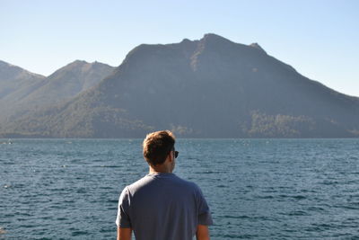 Rear view of man looking at lake and mountains against clear sky
