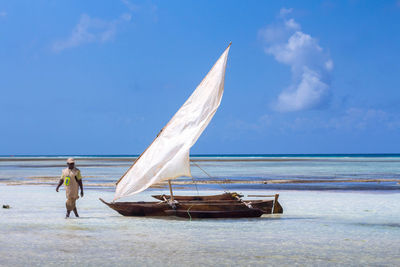 Man walking by sailboat on shore at beach against blue sky