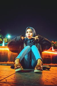 Portrait of young woman wearing hijab while sitting on footpath at night