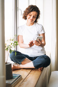 Young brunette woman in casual clothes using mobile phone sitting on window sill at home