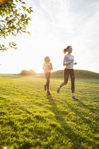 Mother and daughter running on grass at park against sky during sunset