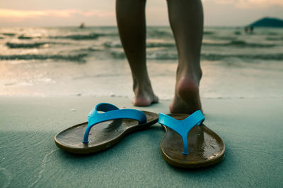 Low section of woman standing on shore with flip-flops in foreground at beach