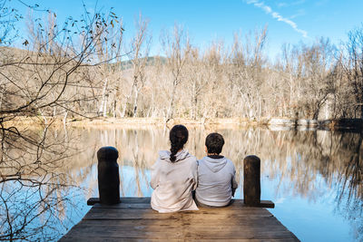 Rear view of people sitting on pier over lake against sky