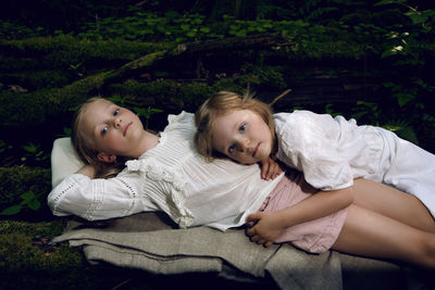 Two girls sisters in white dresses lie on logs in the moss in the forest