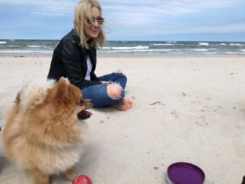 Side view of woman with dog sitting on sand at beach against sky