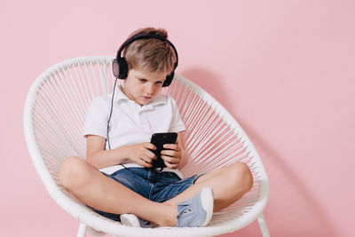 Boy listening music while using phone on chair against colored background