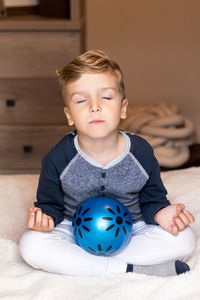 Cute boy with ball sitting on bed at home