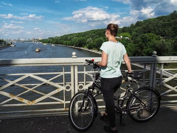 Woman with bicycle on railing by river against sky