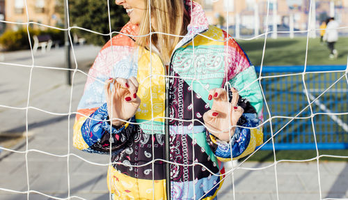 Hands of a young woman clutching the fence on a basketball court wearing a multicolored jacket