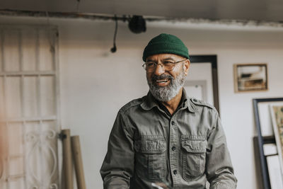 Happy bearded craftsman wearing eyeglasses and knit hat in workshop