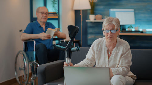 Senior woman using laptop while man reading book on wheelchair