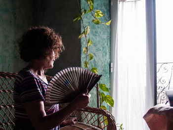 Woman holding hand fan while sitting on seat at home