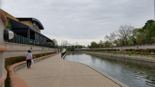 Rear view of man walking on footpath by canal against sky