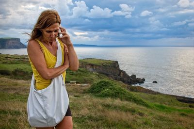 Senior woman standing on field by sea against cloudy sky