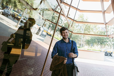 Smiling young businessman with blazer and bag standing outside cafe