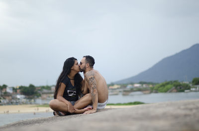 Couple kissing while sitting on beach against sky
