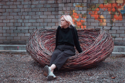 Full length of thoughtful teenage girl sitting in wicker basket against brick wall