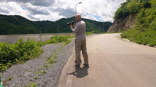 Full length of man holding camera while standing by field on road