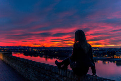 Rear view of woman sitting by sea against sky during sunset