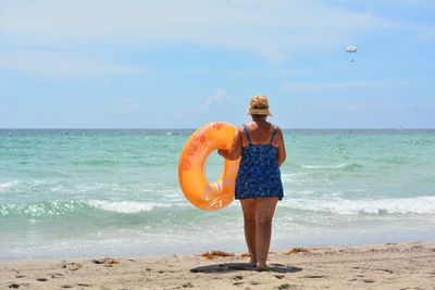 Rear view of woman with inflatable ring walking at beach against sky during sunny day