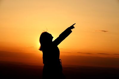Silhouette woman pointing while standing against orange sky