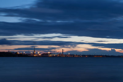Illuminated city at waterfront against cloudy sky