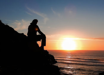 Silhouette man on rock by sea against sky during sunset