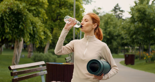 Woman drinking water from bottle while standing in the green park, after fitness training. 