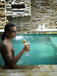 Shirtless young man holding drink while swimming in pool