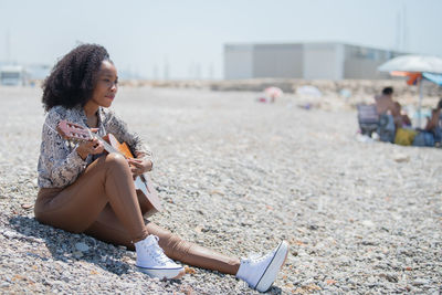 Joyful young woman sitting on the ground of a rocky beach playing her acoustic guitar