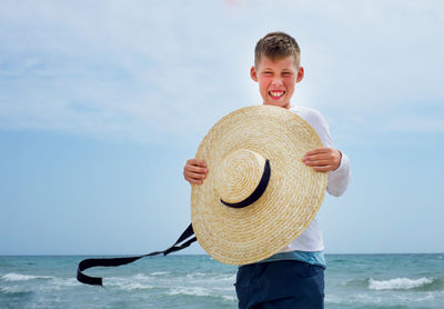 Boy holding hat standing against sea