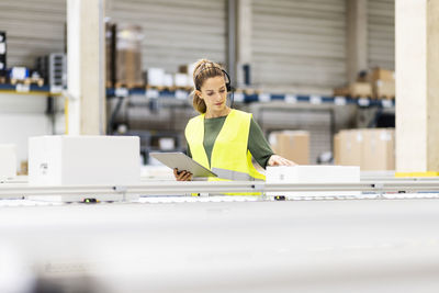 Young worker with tablet pc examining box on conveyor belt in warehouse