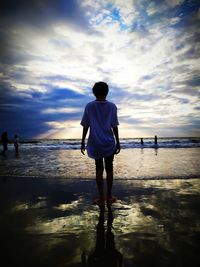 Rear view of man standing at beach against sky during sunset