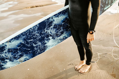 Side view of cropped unrecognizable surfer man dressed in wetsuit standing with the surfboard on the water waiting to catch a wave on the beach during sunrise
