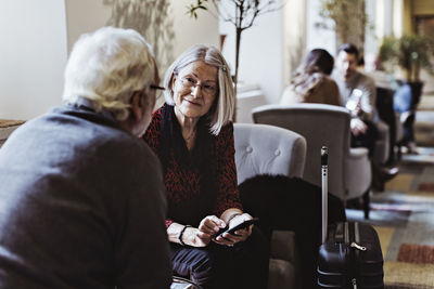 Senior woman with smart phone looking at man while sitting on chair in hotel