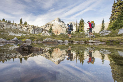 Side view and reflection of backpackers hiking beside alpine lake.