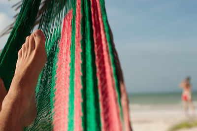 Low section of person relaxing on hammock at beach against sky