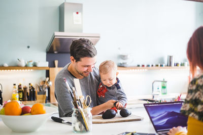 Smiling man carrying son holding tomato on cutting board at kitchen island