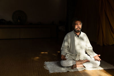 Full body side view of focused bearded ethnic senior male practicing chi kung meditation in lotus position in dark studio