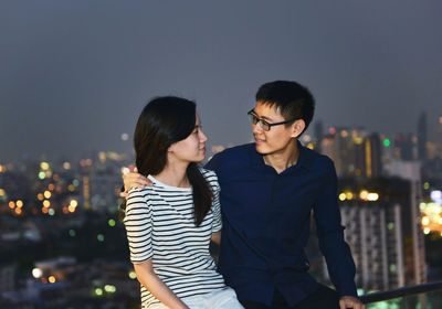 Couple sitting on railing against illuminated city at dusk