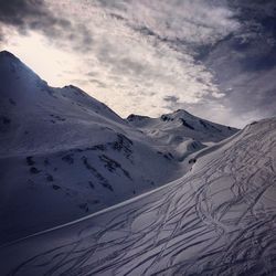 Ski tracks on snowcapped mountains against cloudy sky