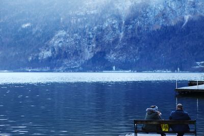 Rear view of friends sitting on bench by lake during winter