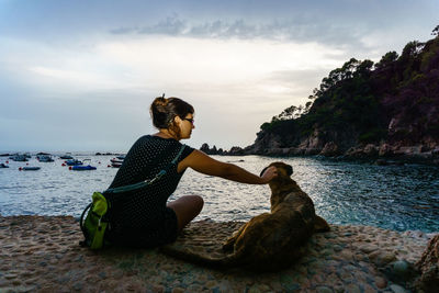 Woman with dog sitting on rock by sea against sky
