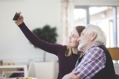 Young woman taking selfie with grandfather while sitting in nursing home